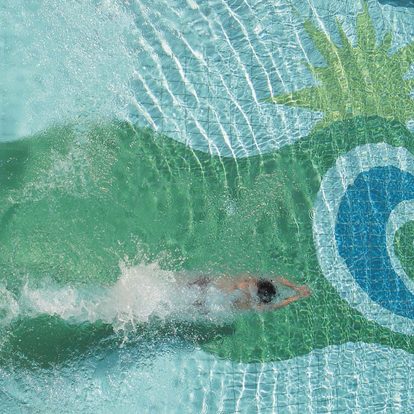 Eine Person schwimmt von oben gesehen durch einen Pool mit Palmen- und Wellenmosaik am Boden.