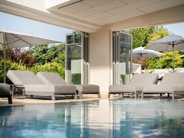 Innenpool mit Liegen, offenen Glastüren, Sonnenschirmen und Gartenblick in einem Wellnessbereich.