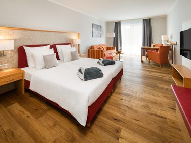 Helles Hotelzimmer mit Doppelbett, Sitzecke, Schreibtisch und Fernseher vor großem Fenster.