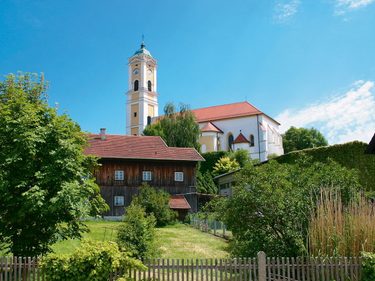 Kirche mit hohem Turm über Garten, Bäumen und Holzhaus in einem Dorf unter blauem Himmel.