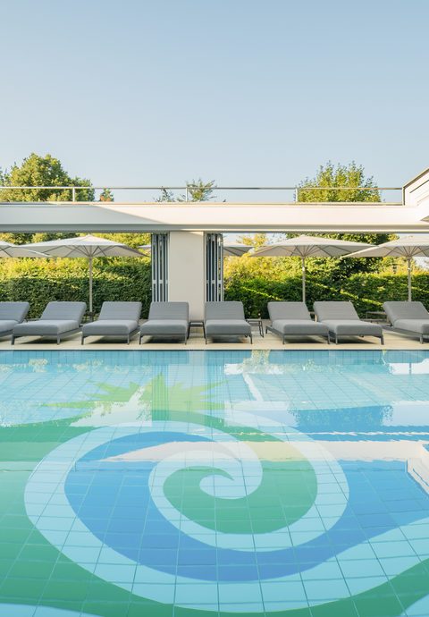 Moderner Innenpool mit Liegen und geöffneten Glasfronten zum begrünten Außenbereich.