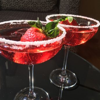 Zwei rote Cocktails mit Zuckerrand, Erdbeere und Minze in Cocktailgläsern auf einem Tisch.