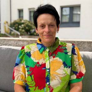 Sitzende Frau in bunter Bluse auf einem Sofa im Außenbereich vor einem Gebäude.