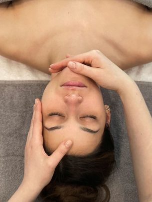 Eine Frau liegt auf einer Wellnessliege und erhält eine Gesichtsbehandlung von zwei Händen.