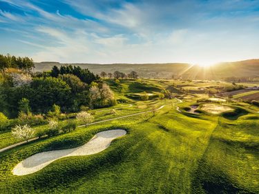Golfplatz mit hügeligen Fairways, Sandbunkern und tief stehender Sonne in weiter Landschaft.