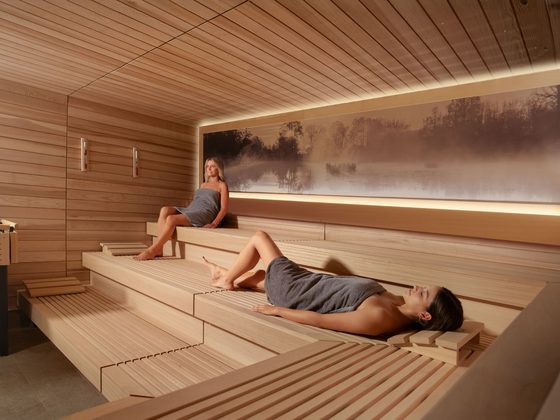 Zwei Frauen mit Handtüchern entspannen auf Holzbänken in einer warm beleuchteten Sauna.