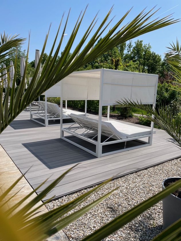 Weiße Daybeds mit Überdachung auf einer Terrasse, umgeben von Palmen, Kies und Bäumen im Garten.