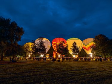 Mehrere beleuchtete Heißluftballons stehen abends auf einer Wiese, umgeben von Besuchern und Bäumen.