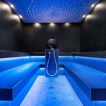 Modernes Dampfbad mit blauer Beleuchtung, schwarzen Wänden, seitlichen Sitzbänken und zentraler Dusche.
