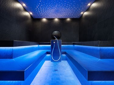 Modernes Dampfbad mit blauer Beleuchtung, schwarzen Wänden, seitlichen Sitzbänken und zentraler Dusche.