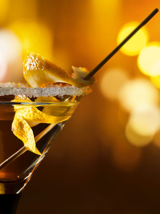 Cocktail im Martiniglas mit Olive, Zitronenzeste und Zuckerrand vor unscharfem Barhintergrund.