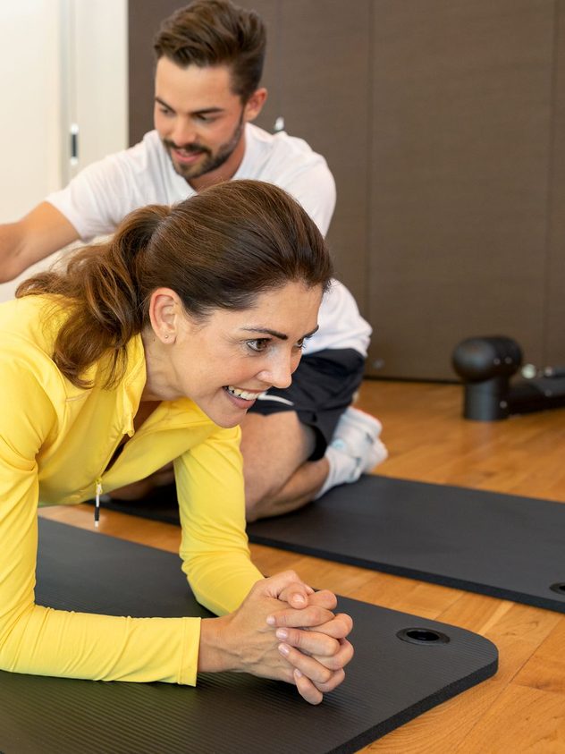 Eine Frau hält eine Plank auf einer Matte, während ein Trainer sie im Fitnessraum anleitet.