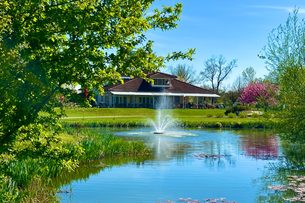 Hotel am Teich mit Springbrunnen, umgeben von Bäumen, Wiese und blauem Himmel.