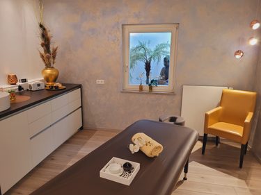 Wellness-Behandlungsraum mit Massageliege, gelbem Sessel und Fenster mit Palme und Buddhafigur.