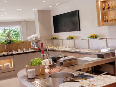 Frühstücksbuffet in einem hellen Restaurant mit offener Kochstation, Buffetauslage und Geschirr.