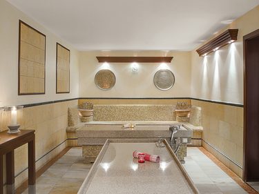 Wellnessraum mit Steinliege, Mosaikbank, zwei Waschbecken und indirekter Beleuchtung.