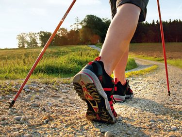 Person beim Nordic Walking auf einem geschwungenen Feldweg in offener Landschaft bei tief stehender Sonne.