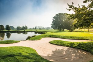 Golfplatz mit Sandbunker, Teich und Bäumen im Morgenlicht.