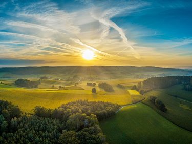 Luftaufnahme von Feldern, Wald und Hügeln bei tief stehender Sonne unter einem wolkigen Himmel.