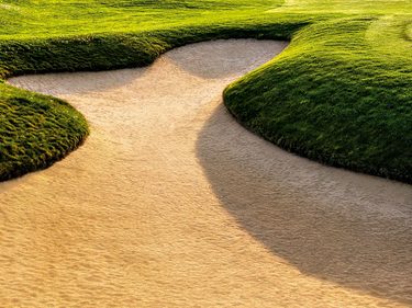 Golfplatz mit geschwungenen Sandbunkern, grünen Hügeln und Bäumen unter blauem Himmel im Abendlicht.