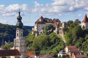 Burganlage und Kirchturm über einer Altstadt mit bunten Häusern auf einem bewaldeten Hügel unter Wolken.