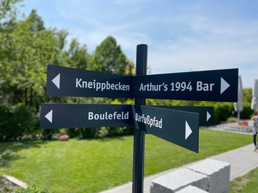 Schwarzer Wegweiser im Hotelgarten mit Richtungen zu Kneippbecken, Bar, Boulefeld und Barfußpfad.