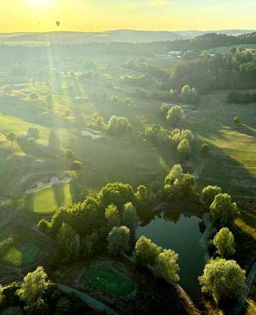 Luftaufnahme einer Hügellandschaft mit Golfplatz, Teich, Bäumen und tief stehender Sonne.