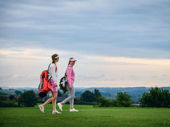 Zwei Golferinnen gehen mit Golftaschen über einen Golfplatz vor weiter Landschaft und wolkigem Himmel.