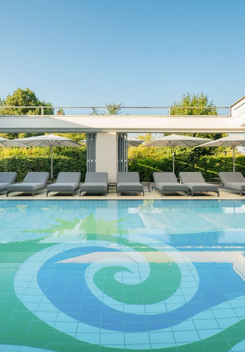 Moderner Innenpool mit Liegen, Glasfronten und Blick auf Hecken und Sonnenschirme im Wellnessbereich.