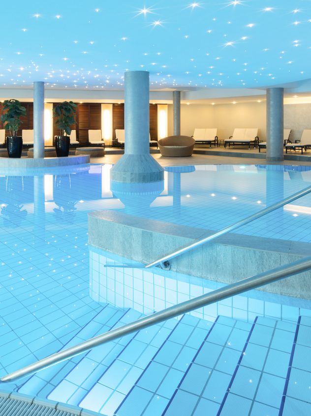 Großer Innenpool im Wellnessbereich mit Treppeneinstieg, Liegen, Säulen und beleuchteter Decke.