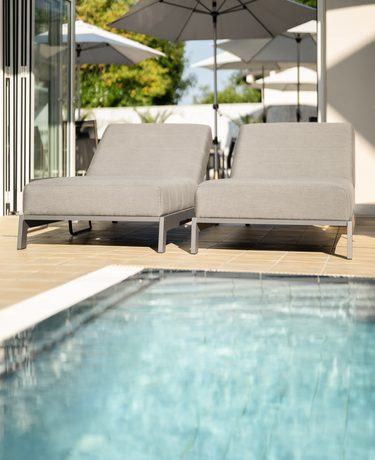 Zwei Liegen auf einer Terrasse am Pool, dahinter mehrere Sonnenschirme und ein offener Übergang zum Außenbereich.