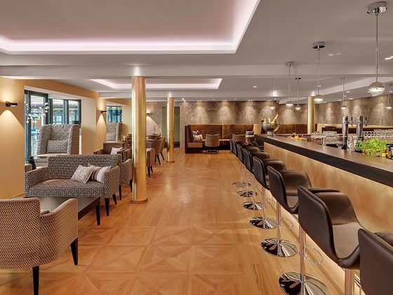 Moderne Hotelbar mit langem Tresen, Barhockern und Sitzgruppen in einem warm beleuchteten Innenraum.