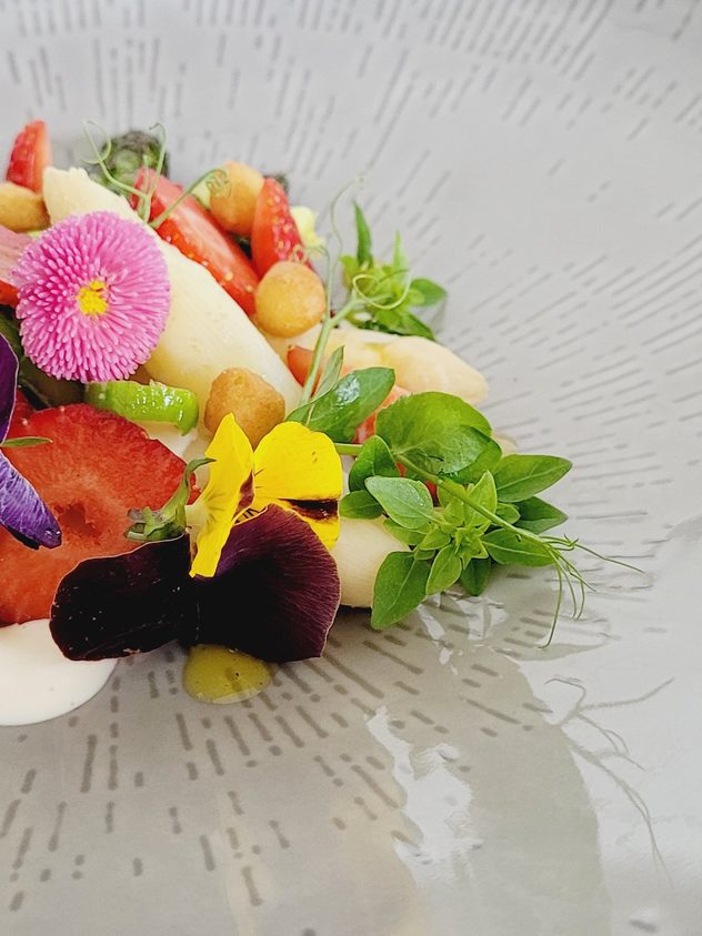 Kreativ angerichtetes Gourmetgericht mit Spargel, Erdbeeren, Kräutern und essbaren Blüten auf einem großen Teller.