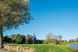 Ein Golfer beim Abschlag auf einem Grün neben einem See unter blauem Himmel.