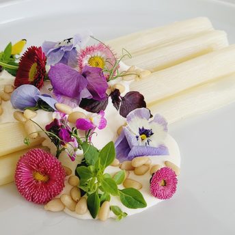 Weißer Spargel mit Creme, Pinienkernen, Kräutern und essbaren Blüten auf einem weißen Teller.