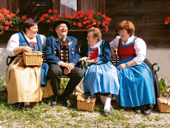 Vier Menschen in Tracht sitzen lachend auf einer Bank vor einem Holzhaus mit roten Geranien.