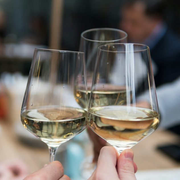 Drei Weingläser mit Weißwein beim Anstoßen im Restaurant, gehalten von Gästen.