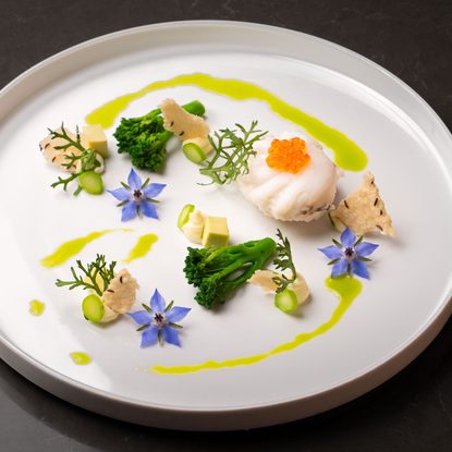 Kreativ angerichtetes Fischgericht mit grünem Gemüse, Kaviar und essbaren Blüten auf weißem Teller.