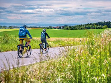 Zwei Radfahrer auf einer Landstraße vor grünen Feldern und bewölktem Himmel.