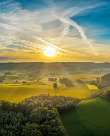 Luftaufnahme von Feldern, Wald und Hügeln bei Sonnenaufgang unter einem Himmel mit Wolken und Kondensstreifen.