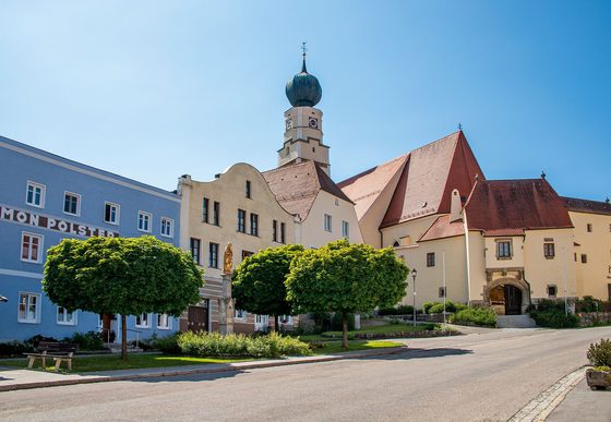 Historische Kirche mit Zwiebelturm an einem sonnigen Ortsplatz, flankiert von Bäumen und Gebäuden.