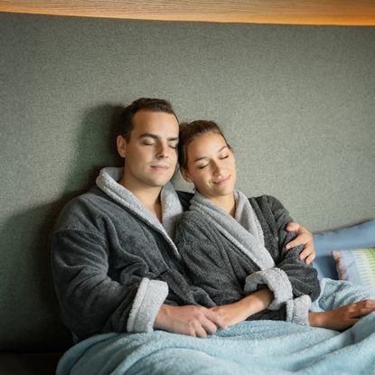 Ein Paar sitzt in Bademänteln unter einer Decke in einem Wellness-Ruheraum und entspannt.