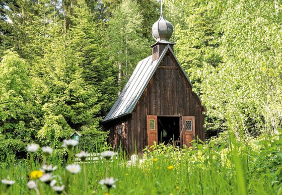 Kleine hölzerne Kapelle in einer Wiese mit Blumen, umgeben von Bäumen und Wald.