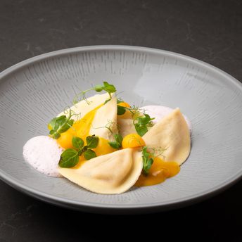 Drei Ravioli in einer grauen Schale mit gelber Sauce, hellem Schaum und Microgreens.