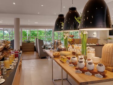 Frühstücksbuffet in einem hellen Restaurant mit Speisen, Getränken, Tischen und großen Fenstern.