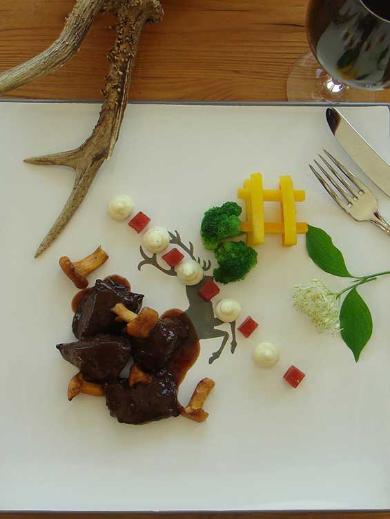 Kunstvoll angerichtetes Wildgericht auf einem weißen Teller mit stilisiertem Hirschmotiv. Das Gericht besteht aus dunklem Fleisch mit Pilzen in Sauce, kleinen Gemüsewürfeln, weißen Cremetupfen, Brokkoliröschen und dekorativ angeordneten Pommes frites. Der Teller ist mit Holzelementen, Blättern, Blüten und einem Hirschgeweih dekoriert. Im Hintergrund liegt Besteck und ein Weinglas auf einem rustikalen Holztisch