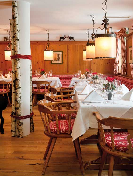 Gemütlich eingerichtete „Hirschstube“ im Wellnesshotel Sonnengut mit liebevoll gedeckten Tischen, Holzvertäfelung, traditionellen Stühlen und roter Alpen-Deko. Weiß eingedeckte Tafeln mit Blumen und Gläsern schaffen eine elegante, warme Atmosphäre.