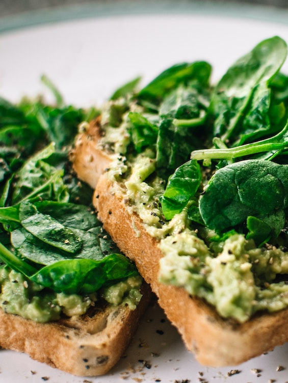Zwei Scheiben Vollkorntoast mit cremigem Avocado-Aufstrich, belegt mit frischem Spinat und gewürzt mit schwarzem Pfeffer und anderen Kräutern. Die Nahaufnahme zeigt die Textur der Zutaten auf einem weißen Teller.