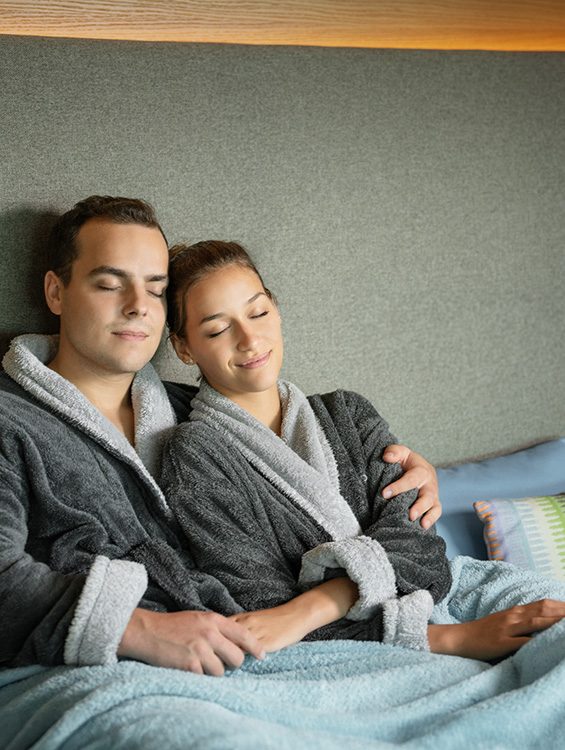 Ein lächelndes Paar in kuscheligen, grauen Bademänteln sitzt entspannt auf einem Sofa. Der Mann hat seinen Arm um die Frau gelegt, beide wirken glücklich und genießen den Moment.