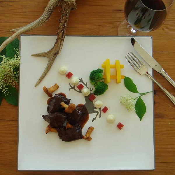 Kunstvoll angerichtetes Wildgericht auf einem weißen Teller mit stilisiertem Hirschmotiv. Das Gericht besteht aus dunklem Fleisch mit Pilzen in Sauce, kleinen Gemüsewürfeln, weißen Cremetupfen, Brokkoliröschen und dekorativ angeordneten Pommes frites. Der Teller ist mit Holzelementen, Blättern, Blüten und einem Hirschgeweih dekoriert. Im Hintergrund liegt Besteck und ein Weinglas auf einem rustikalen Holztisch